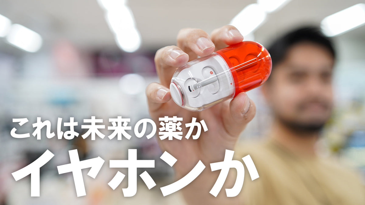 赤と透明な素材で作られたカプセル型のユニークなワイヤレスイヤホン充電ケースを、男性が手に持ってカメラに向けている。背景は店舗内のようにぼかされている。画像下部には「これは未来の薬か」「イヤホンか」という大きなキャッチコピーが書かれている。
